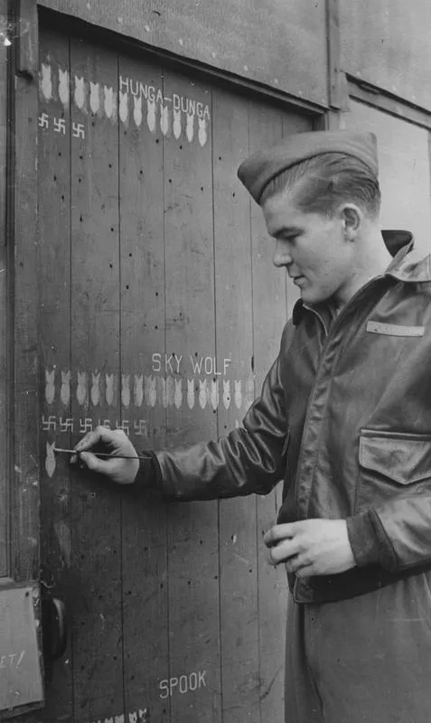 Staff Sergeant Albert Van Hoon Carroll, a gunner of the 303rd Bomb Group marks raids on the door to an army hut. Image stamped on reverse: 'Copyright Current Affairs Ltd.' [stamp], 'Passed for Publication 9 Apr 1943.' [stamp] and '257542.' [Censor no.] Printed caption on reverse: S/Sgt. (Gunner) Albert Van Hoon Carroll, aged 20, of 110 W. Fannin Marshall, Texas, marks up the record of another raid on a door at his English base. Carroll, a student before joining the U.S. Air Force, has taken part in 11 rai